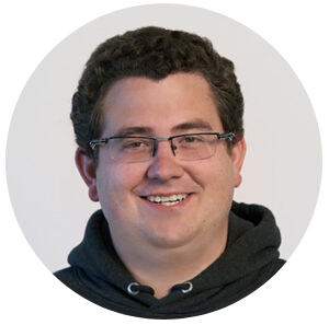 Ein lächelnder Mann mit kurzen lockigen braunen Haaren, der eine Brille und einen dunklen Kapuzenpulli trägt, ist vor einem schlichten, hellen Hintergrund in einem runden Rahmen abgebildet.