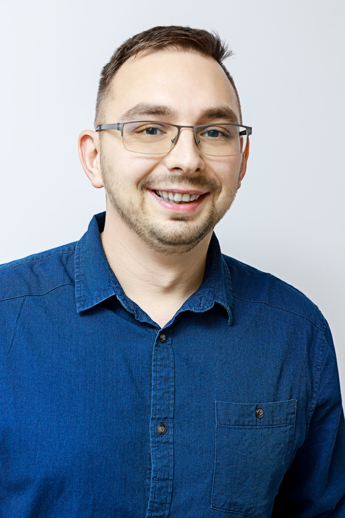 Mężczyzna z krótkimi brązowymi włosami, okularami i przystrzyżoną brodą uśmiecha się do kamery. Ma na sobie niebieską koszulę zapinaną na guziki i stoi na białym tle.