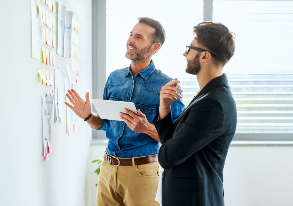 Zwei Männer stehen in einem Büro. Der eine im Jeanshemd hält ein Tablet in der Hand und gestikuliert in Richtung einer Wand, die mit Papieren über VMware Offering für Service Provider bedeckt ist, während der andere Mann im Anzug nachdenklich zuhört.